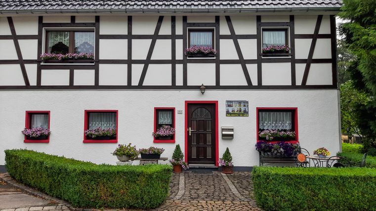 A charming half-timbered house with a white facade and red windows. In front of the house, there are well-maintained plants and a paved path.