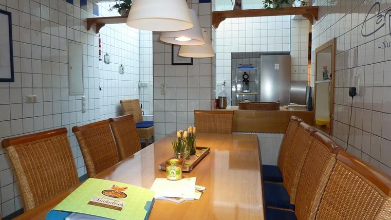 A cozy dining area with a long wooden table and rattan chairs. The walls are tiled, and bright lamps hang above the table.