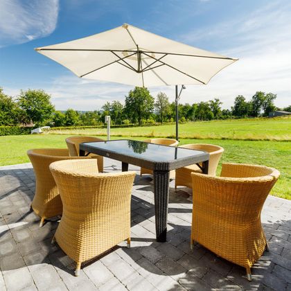 A cozy outdoor dining area with a large table and six wicker chairs. A parasol provides shade from the sun, surrounded by green grass.