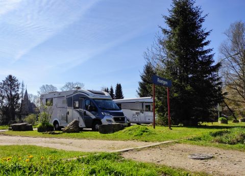 A beautiful campsite with two motorhomes and a green environment. In the background, trees and a path are visible.