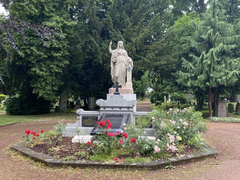 Eine Statue steht inmitten eines freundlich gestalteten Grabes, umgeben von bunten Blumen. Im Hintergrund sind hohe Bäume sichtbar, die eine ruhige Atmosphäre schaffen.