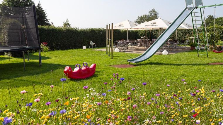 A beautiful garden with colorful flowers and a slide. In the background, there is a trampoline and tables under sun umbrellas.
