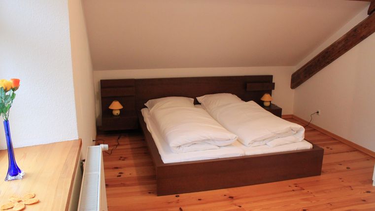 Een gezellige slaapkamer met een tweepersoonsbed en een houten vloer. Op het nachtkastje staan twee lampen en een vaas met bloemen.