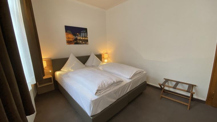 A cozy bedroom with two single beds and warm light. A picture hangs on the wall, and there is a small stool for additional comfort.