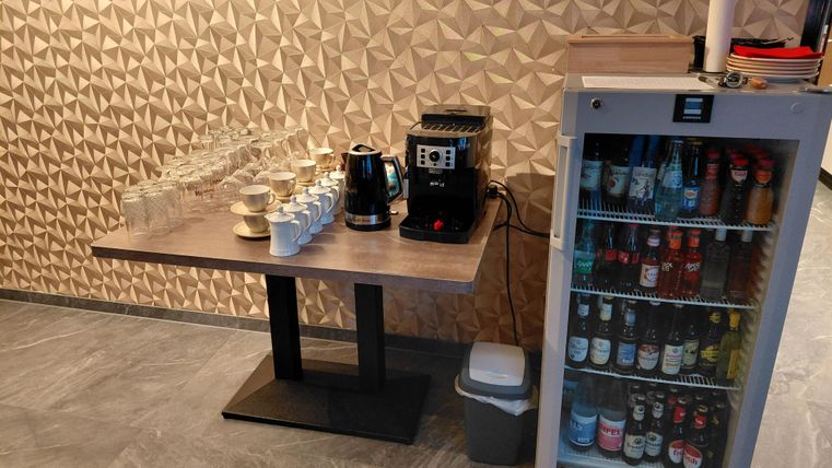 A coffee station with glasses and a coffee machine is on a table. Next to it is a refrigerator with various drinks.