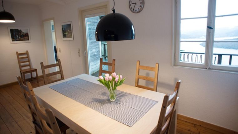 A cozy dining area with a large table and wooden chairs. On the table, there is a vase with flowers and the room has large windows overlooking the landscape.