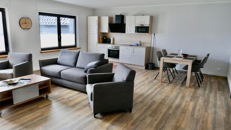 A modern living room with a gray sofa and a wooden table. The open kitchen corner is well-equipped and light pours in through the large windows.